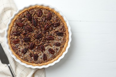 Delicious pecan pie in baking dish and knife on white wooden table, flat lay. Space for text