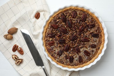 Photo of Delicious pecan pie in baking dish, knife and fresh nuts on white wooden table, flat lay