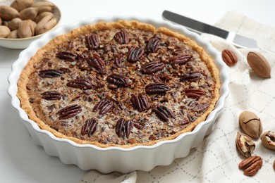 Photo of Delicious pecan pie in baking dish and fresh nuts on white wooden table, closeup