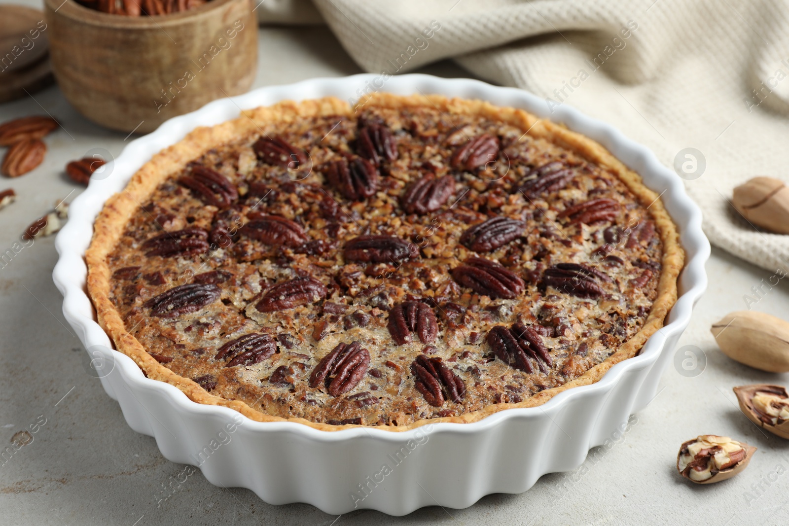 Photo of Delicious pecan pie in baking dish and fresh nuts on gray textured table, closeup