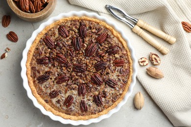 Photo of Delicious pecan pie in baking dish, nutcracker and fresh nuts on gray textured table, flat lay