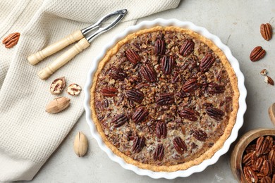 Photo of Delicious pecan pie in baking dish, nutcracker and fresh nuts on gray textured table, flat lay