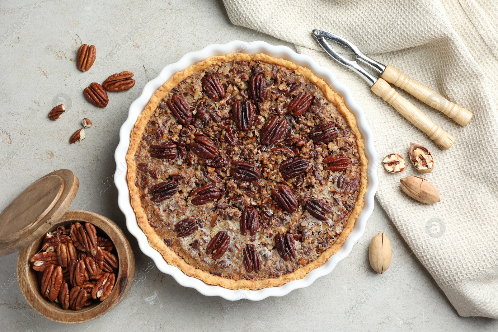 Photo of Delicious pecan pie in baking dish, nutcracker and fresh nuts on gray textured table, flat lay