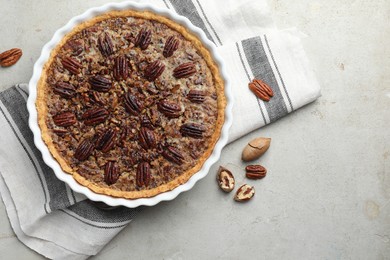 Photo of Delicious pecan pie in baking dish and fresh nuts on gray textured table, top view