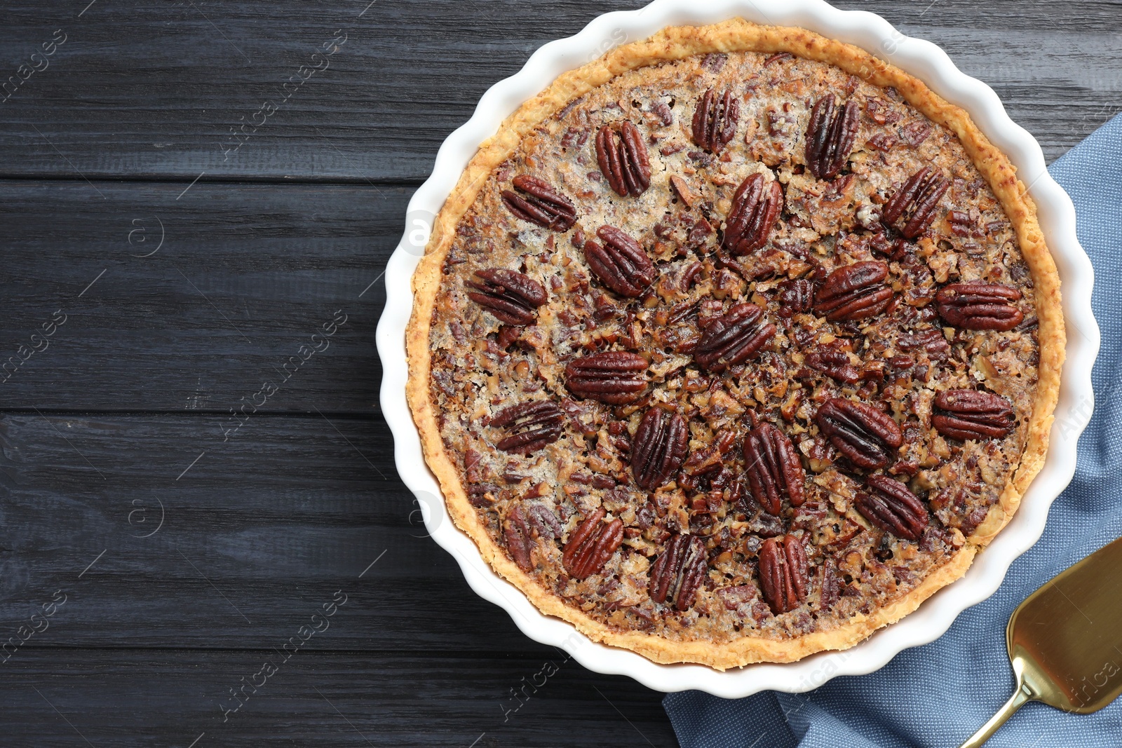 Photo of Delicious pecan pie in baking dish and cake server on dark wooden table, flat lay. Space for text