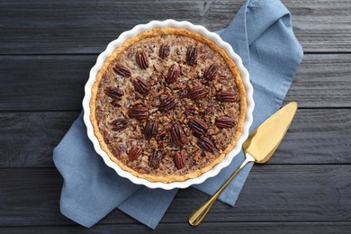 Delicious pecan pie in baking dish and cake server on dark wooden table, flat lay