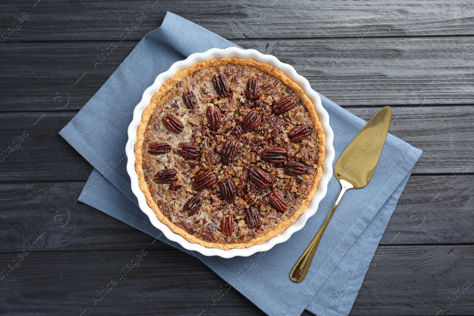 Photo of Delicious pecan pie in baking dish and cake server on dark wooden table, flat lay