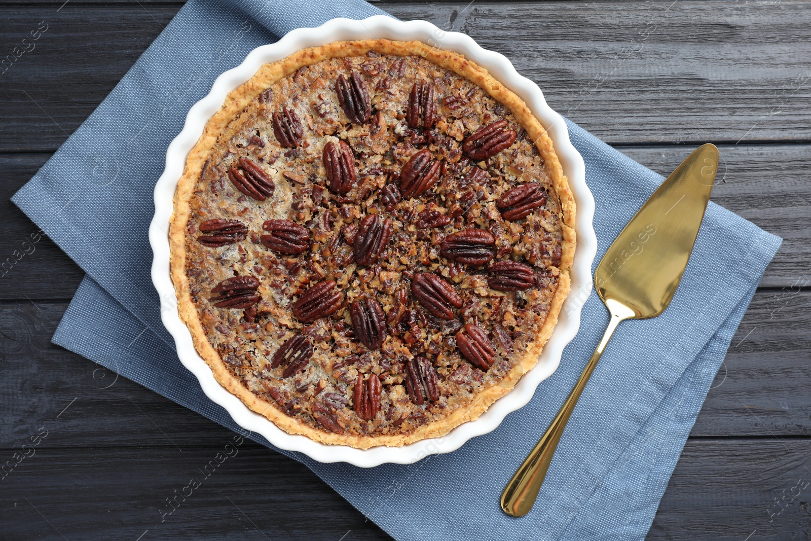 Photo of Delicious pecan pie in baking dish and cake server on dark wooden table, flat lay