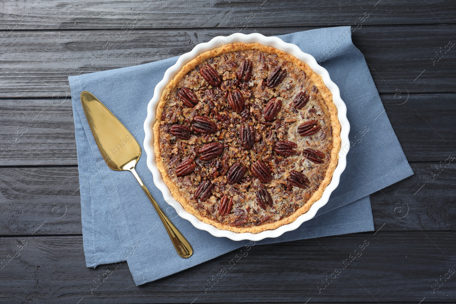 Photo of Delicious pecan pie in baking dish and cake server on dark wooden table, flat lay