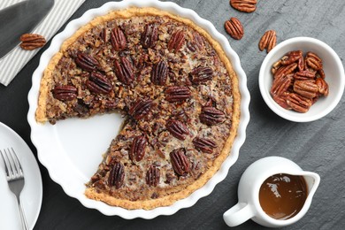 Photo of Delicious pecan pie, fresh nuts, caramel sauce and cake server on dark gray textured table, top view