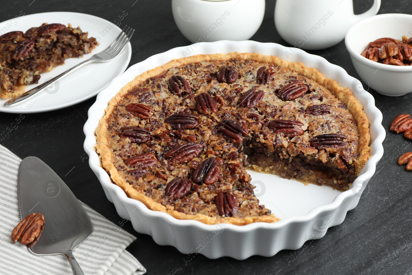 Photo of Delicious pecan pie in baking dish and cake server on dark gray textured table, closeup