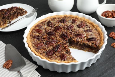 Photo of Delicious pecan pie in baking dish and cake server on dark gray textured table, closeup