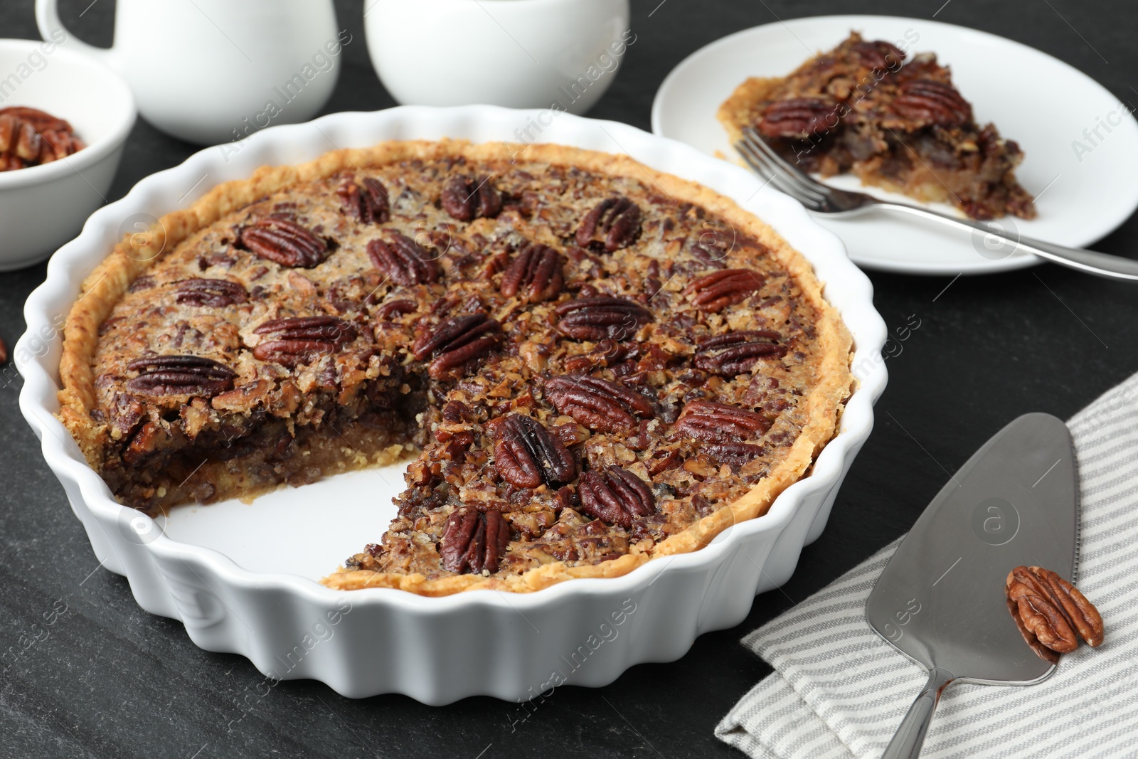 Photo of Delicious pecan pie in baking dish and cake server on dark gray textured table, closeup