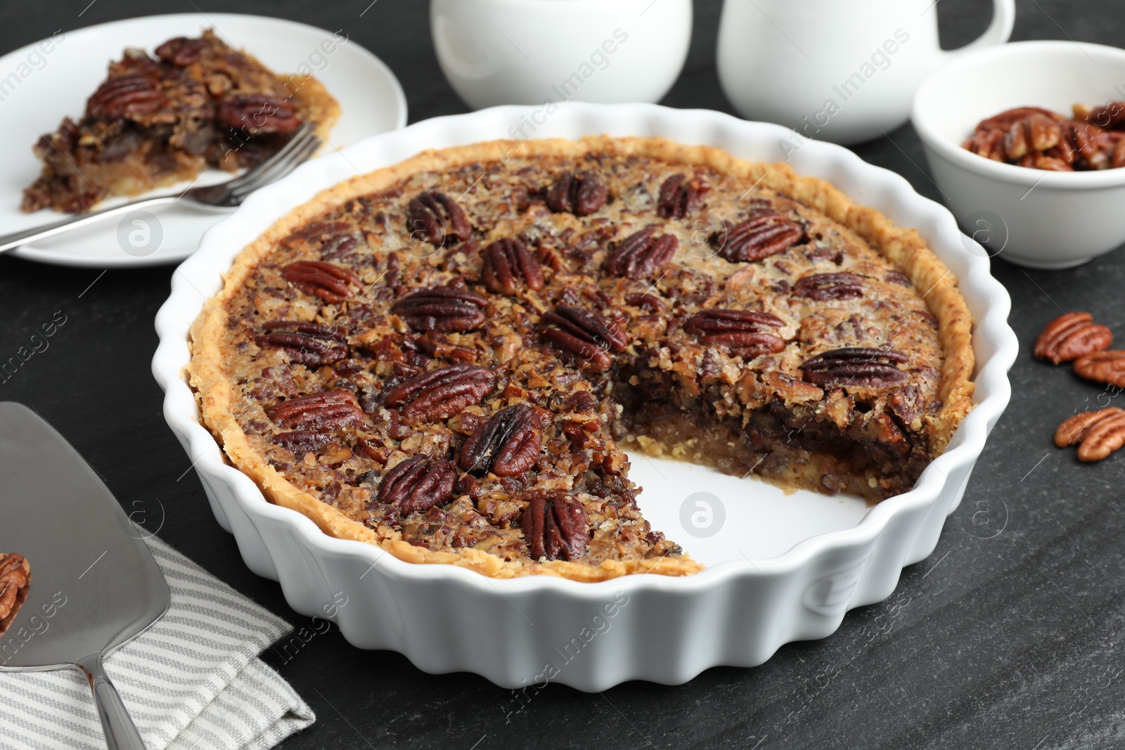 Photo of Delicious pecan pie in baking dish and cake server on dark gray textured table, closeup