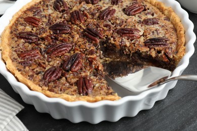 Photo of Delicious pecan pie in baking dish and cake server on dark gray textured table, closeup