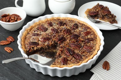 Photo of Delicious pecan pie in baking dish and cake server on dark gray textured table, closeup