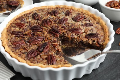 Photo of Delicious pecan pie in baking dish and cake server on dark gray textured table, closeup