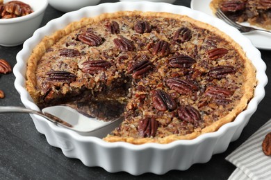 Photo of Delicious pecan pie in baking dish and cake server on dark gray textured table, closeup