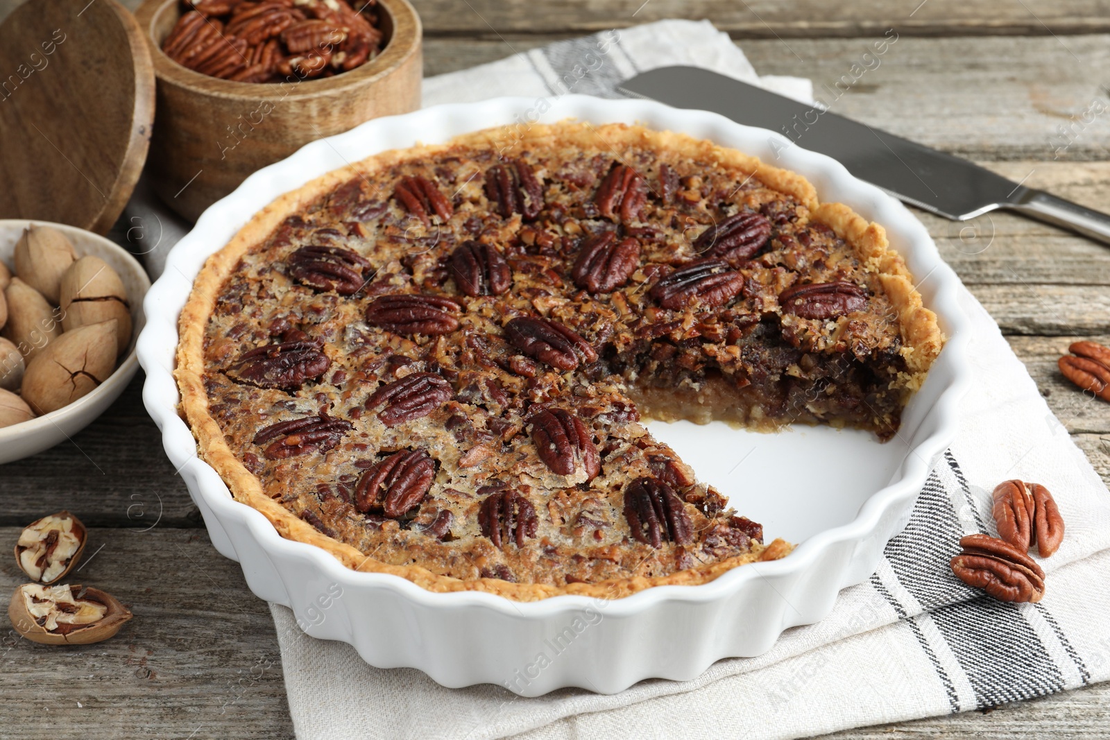 Photo of Delicious pecan pie in baking dish, fresh nuts and knife on wooden table, closeup