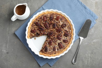 Delicious pecan pie in baking dish, caramel sauce and cake server on gray textured table, flat lay