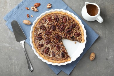 Delicious pecan pie in baking dish, caramel sauce, cake server and fresh nuts on gray textured table, flat lay