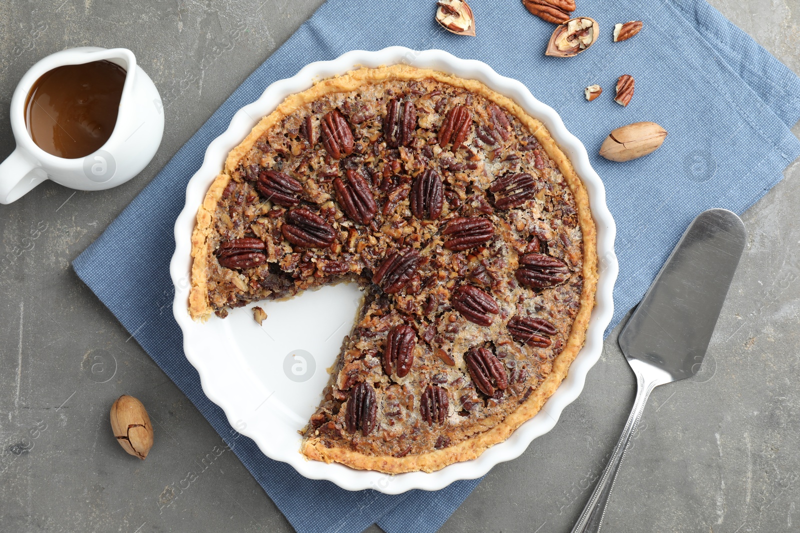 Photo of Delicious pecan pie in baking dish, caramel sauce, cake server and fresh nuts on gray textured table, flat lay