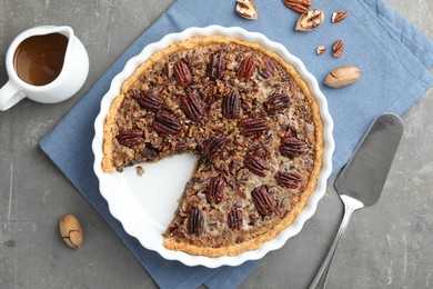 Delicious pecan pie in baking dish, caramel sauce, cake server and fresh nuts on gray textured table, flat lay