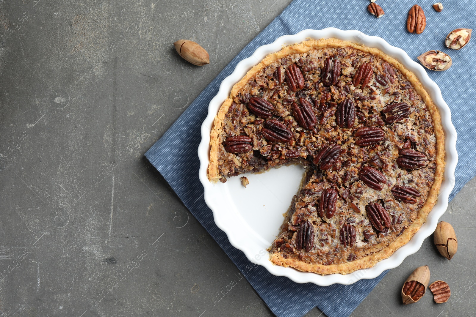 Photo of Delicious pecan pie in baking dish and fresh nuts on gray textured table, top view. Space for text