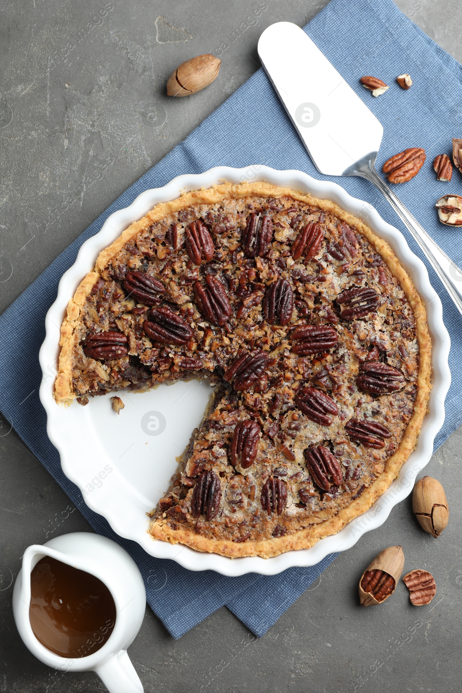 Photo of Delicious pecan pie in baking dish, caramel sauce, cake server and fresh nuts on gray textured table, flat lay