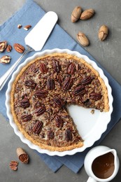 Photo of Delicious pecan pie in baking dish, caramel sauce, cake server and fresh nuts on gray textured table, flat lay