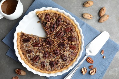 Photo of Delicious pecan pie in baking dish, caramel sauce, cake server and fresh nuts on gray textured table, flat lay