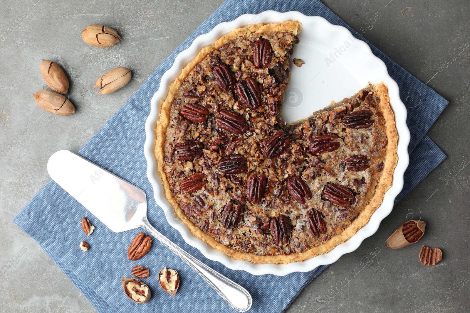 Photo of Delicious pecan pie in baking dish, cake server and fresh nuts on gray textured table, flat lay