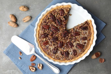 Photo of Delicious pecan pie in baking dish, cake server and fresh nuts on gray textured table, flat lay