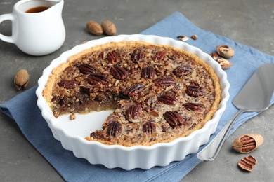 Photo of Delicious pecan pie in baking dish, cake server and fresh nuts on gray textured table, closeup