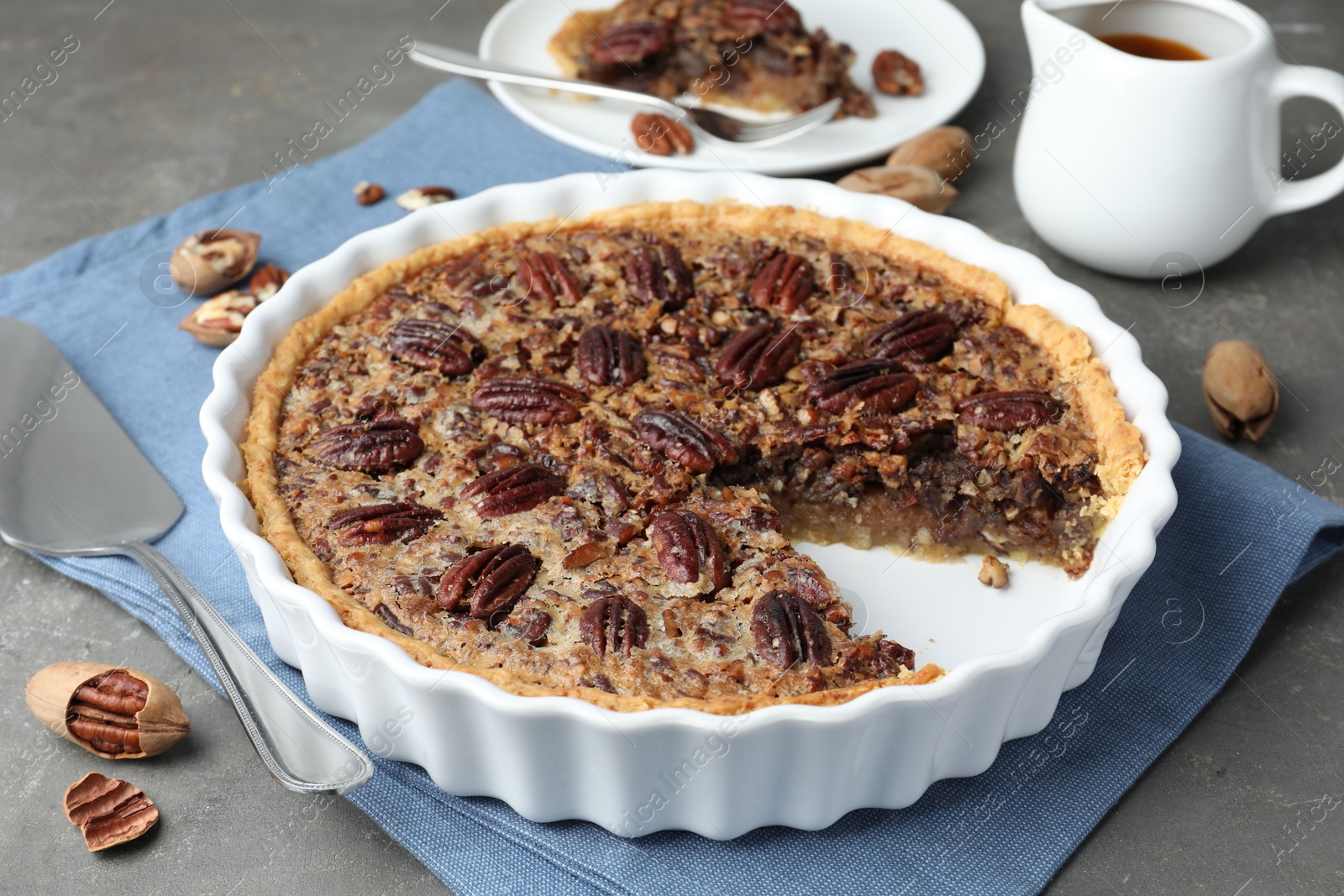 Photo of Delicious pecan pie in baking dish, cake server and fresh nuts on gray textured table, closeup