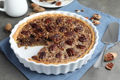 Photo of Delicious pecan pie in baking dish, cake server and fresh nuts on gray textured table, closeup