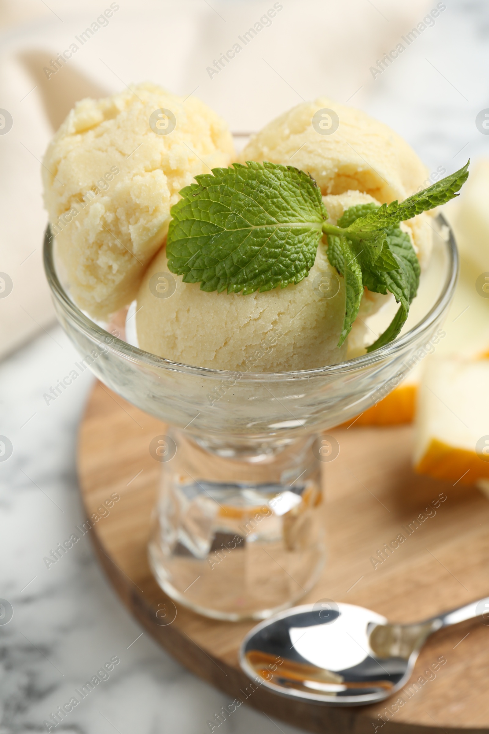 Photo of Scoops of melon sorbet with mint in glass dessert bowl, fresh fruit and spoon on light marble table, closeup