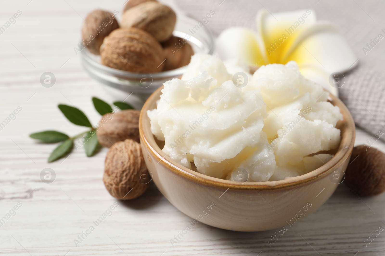 Photo of Shea butter in bowl, flower and nuts on white wooden table, closeup