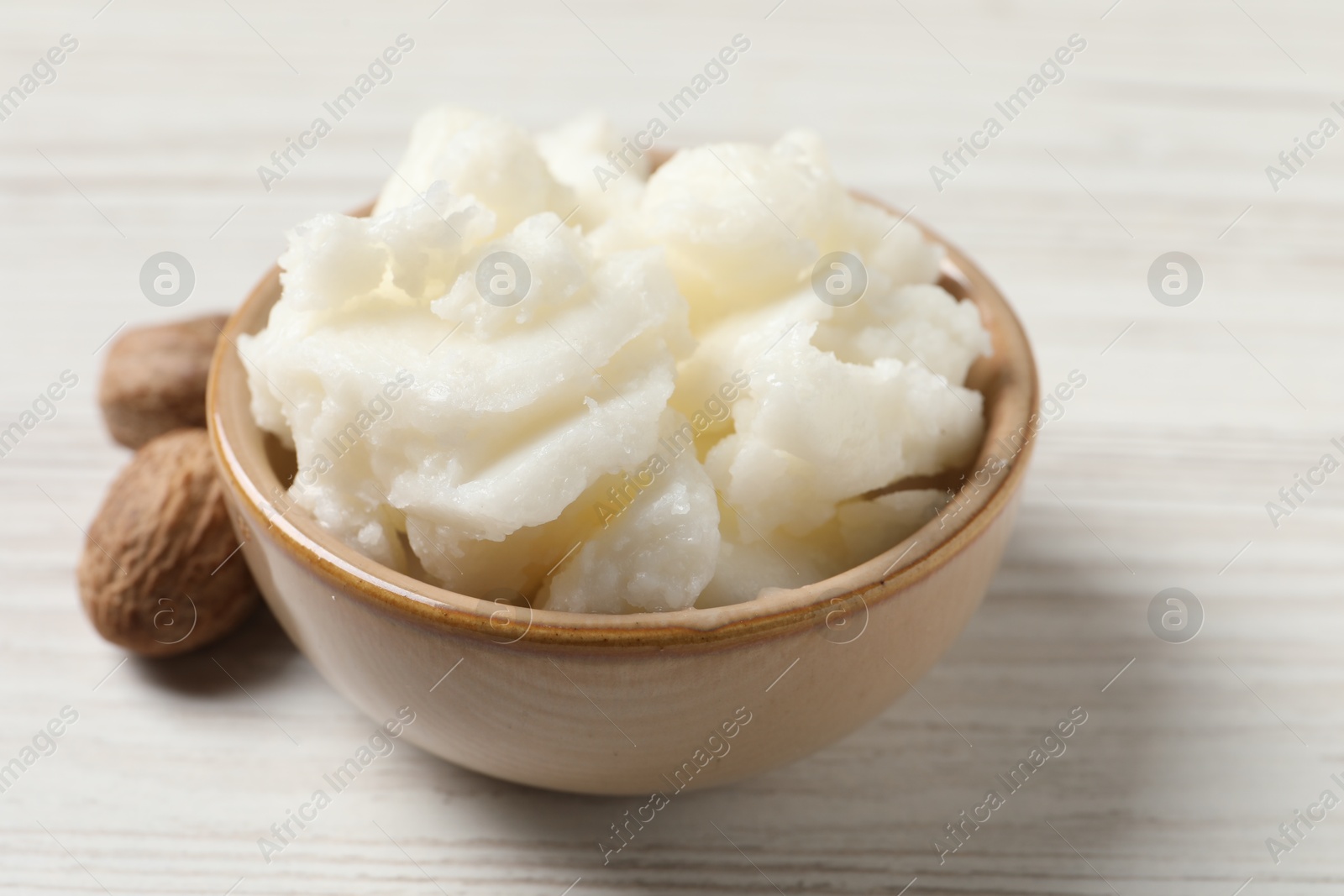 Photo of Shea butter in bowl and nuts on white wooden table, closeup