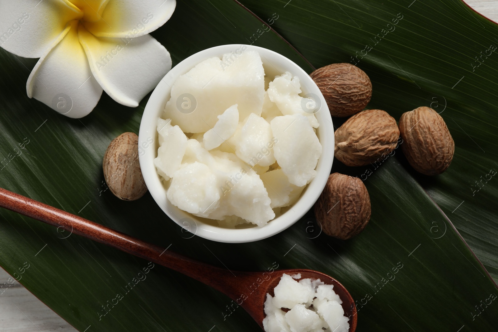 Photo of Shea butter in bowl, spoon, flower and nuts on white wooden table, flat lay