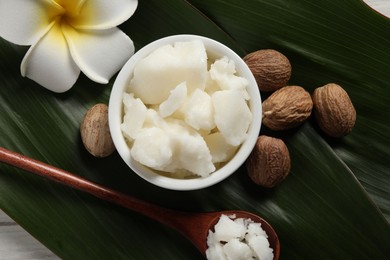 Photo of Shea butter in bowl, spoon, flower and nuts on white wooden table, flat lay