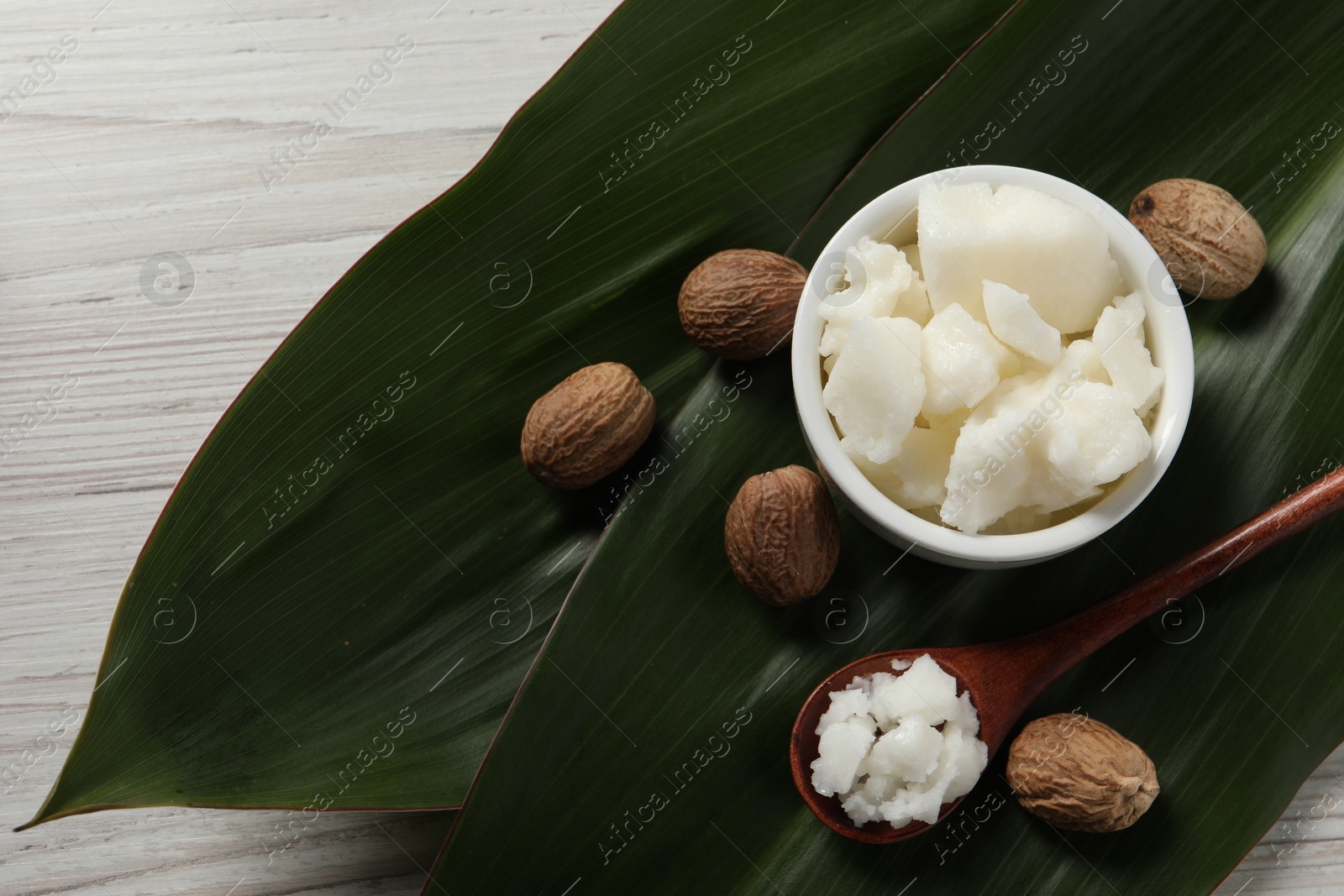 Photo of Shea butter in bowl, spoon, and nuts on white wooden table, flat lay. Space for text