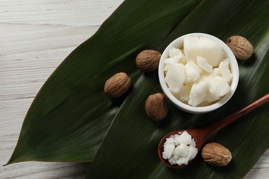 Shea butter in bowl, spoon, and nuts on white wooden table, flat lay. Space for text