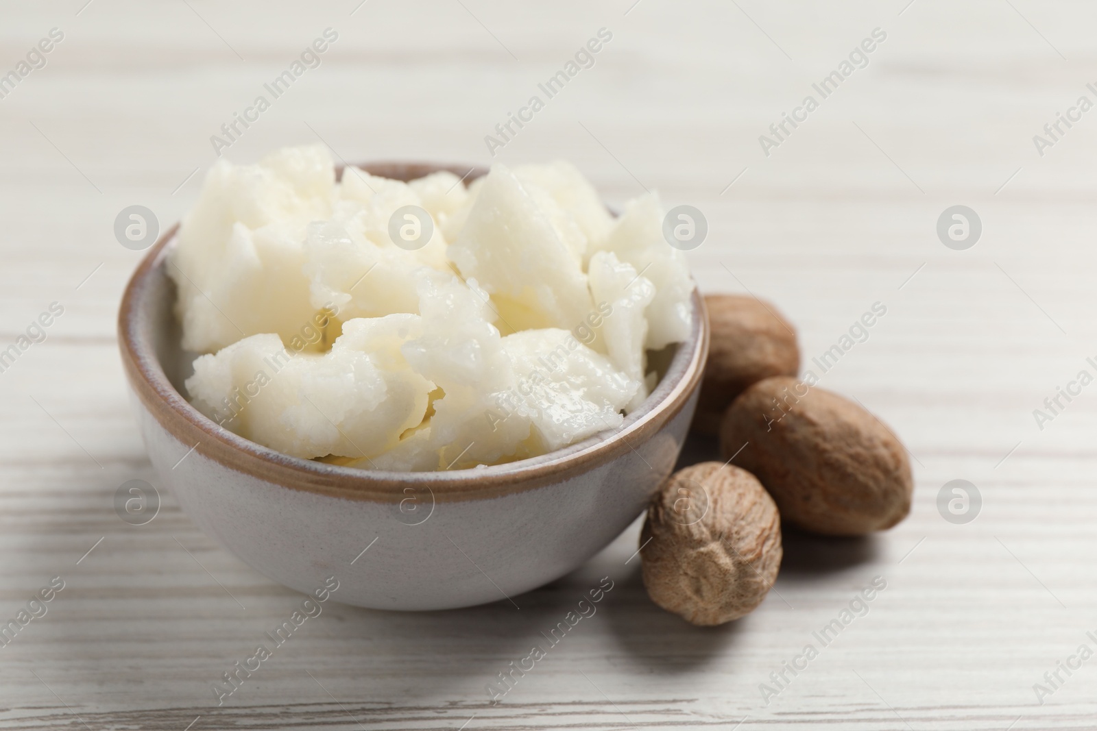 Photo of Shea butter in bowl and nuts on white wooden table, closeup