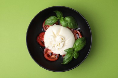 Photo of Delicious burrata cheese, tomatoes and basil in bowl on green table, top view