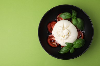 Photo of Delicious burrata cheese, tomatoes and basil in bowl on green table, top view. Space for text