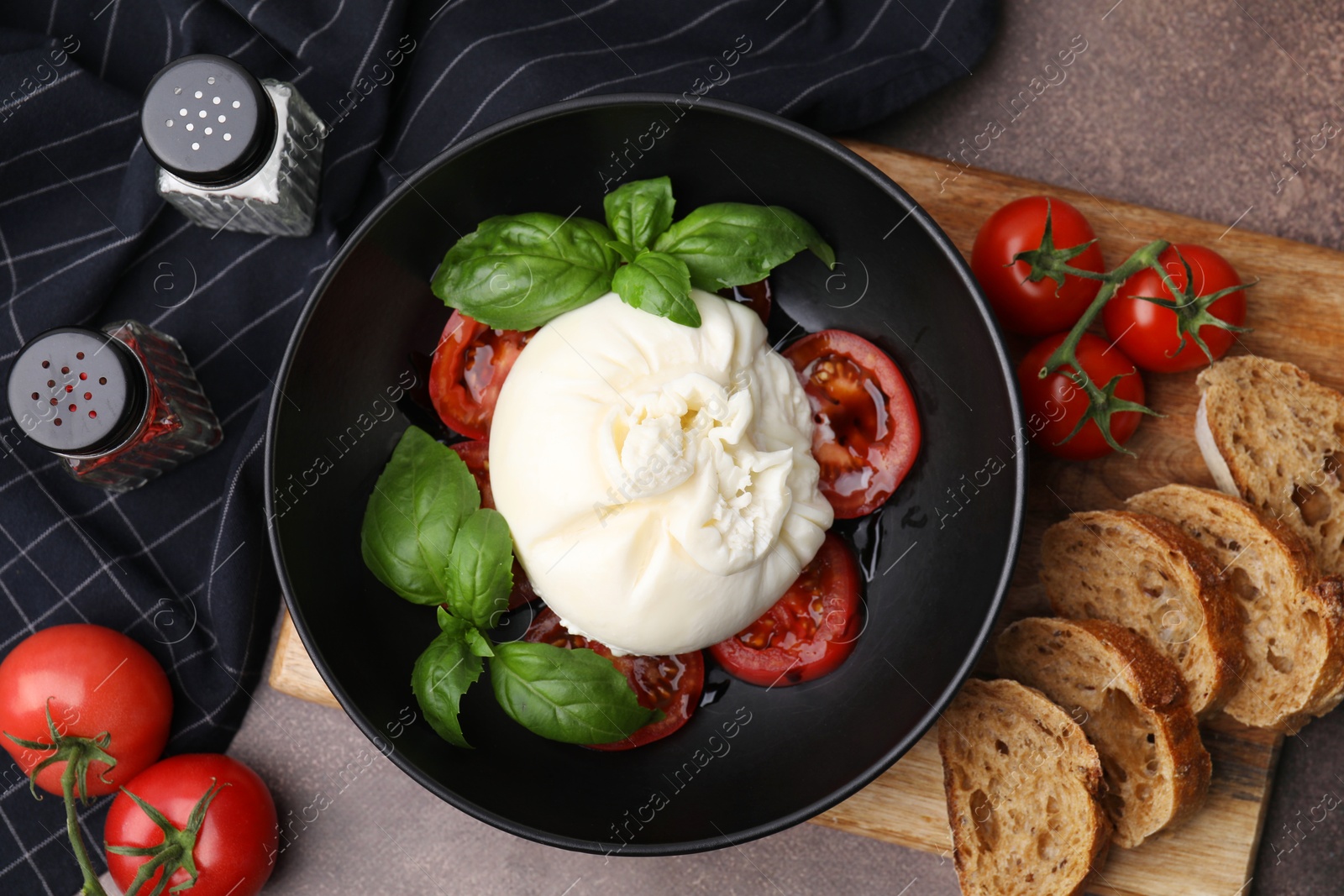 Photo of Delicious burrata cheese, tomatoes, basil and bread in bowl on brown table, flat lay