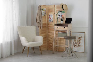 Photo of Folding screen, table, laptop, lamp and armchair indoors. Interior design