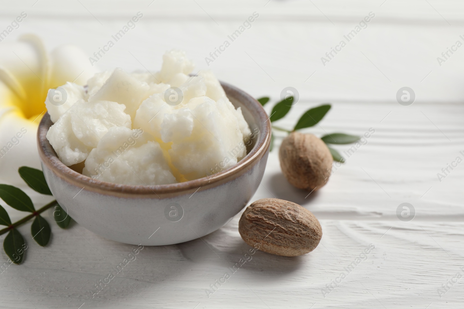 Photo of Shea butter in bowl, flower and nuts on white wooden table, closeup