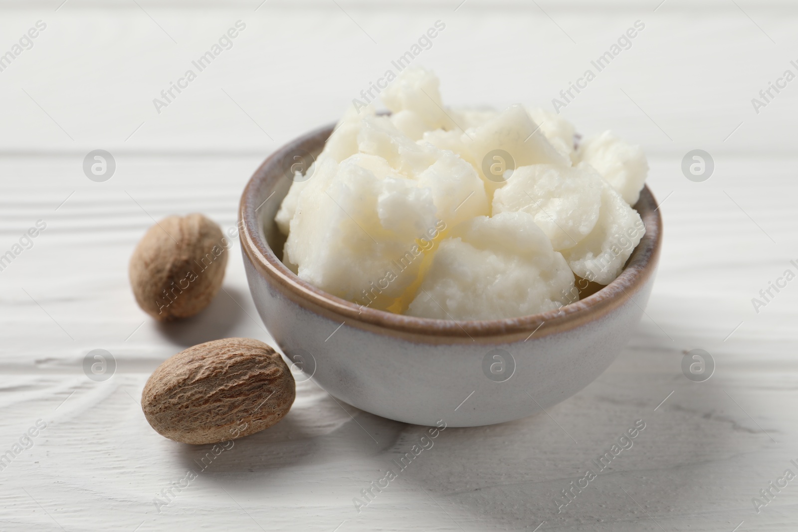 Photo of Shea butter in bowl and nuts on white wooden table, closeup